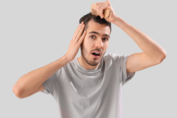 Shocked young man with hair loss problem combing on light background