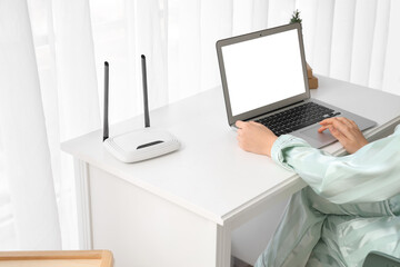 Woman with wi-fi router using laptop on table at home, closeup