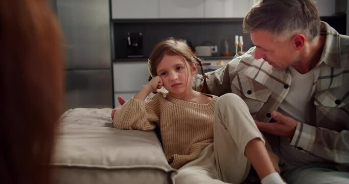 Sad and indignant little girl sits on the sofa and listens to her parents during general communication on the sofa in the evening. Parents punish a little girl and tell her why she is wrong in a