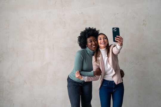 Two happy carefree women enjoying themselves while standing together and taking selfies with smart phone.