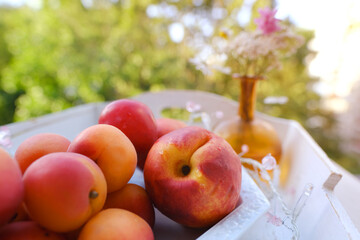 on wooden white tray plate with orange apricots, peach, bunch wild flowers on table in garden, beautiful summer still life, fruits, green trees in background, concept picnic in nature, healthy eating