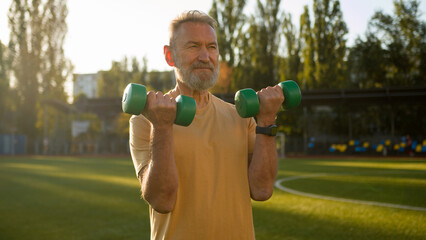 Old Caucasian man grandfather lift dumbbell pump biceps sport athlete stadium outside sportsman...