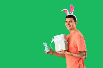 Happy young man in bunny ears with gift box and toy rabbit on green background. Easter celebration