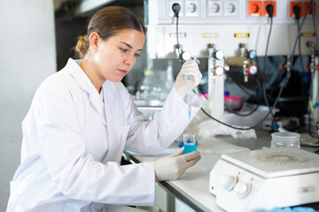 Focused female chemist pipetting blue reagent into solution in test tube while performing...