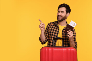 Smiling man with passport, tickets and suitcase pointing at something on yellow background. Space for text