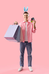 Young man in bunny ears with shopping bags and credit cards on pink background. Easter Sale