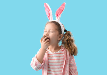 Cute little girl in bunny ears eating chocolate Easter egg on blue background