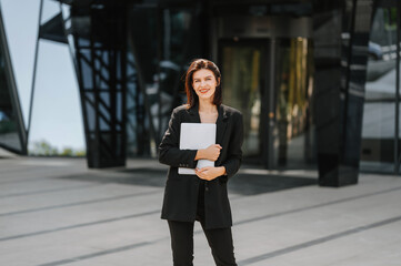 Cheerful young successful woman wearing suit carrying a laptop on a sunny day near a business building.