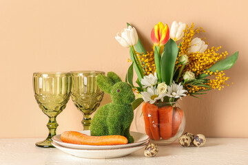 Beautiful table setting for Easter decorated with bouquet of carrots and flowers near beige wall