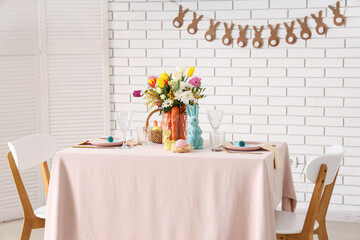 Beautiful table setting for Easter decorated with bouquet of carrots and flowers near white brick wall
