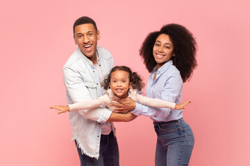 Family fun. Happy black family playing with their little daughter, cute girl pretending flying, spreading hands, imitating plane, posing on pink background