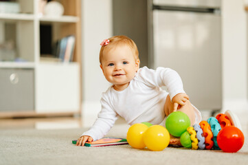 Cheerful baby girl is playing with colourful toys while sitting on the floor at home.