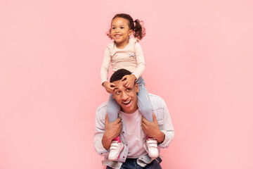 Happy family moments. Cute black little girl sitting on father's shoulders, posing together and having fun on pink background, copy space