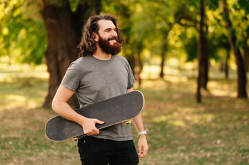 Smiling bearded man with long hair is holding his skateboard while walking in park. © Vulp