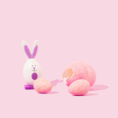 The rabbit stands by the Easter eggs. Biggest egg crack. Pastel pink background. Minimal holiday Easter concept. Copy space. Frontal view. Monochromatic colors.