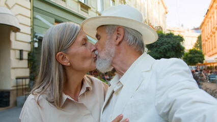 Happy elderly Caucasian tourists couple senior man woman taking selfie streaming video romantic kissing mobile phone modern technologies outside city street. Social media holidays vacations retirement