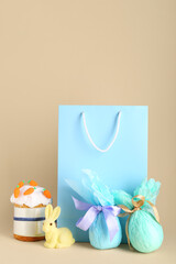 Shopping bag with Easter cake, wrapped eggs and bunny figurine on beige background