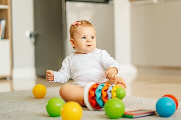 Cute sitting on the floor little baby girl is playing with colourful toys at home.