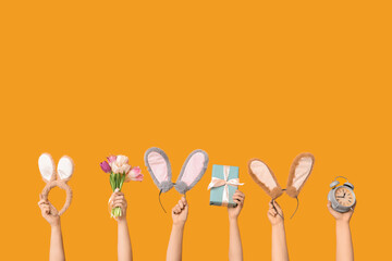 Female hands holding Easter bunny ears headbands with gift box, alarm clock and tulips on orange...