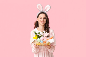 Happy young woman in bunny ears headband holding basket with flowers, Easter eggs and carrot on pink background