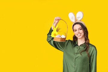 Happy young woman in bunny ears headband holding basket with flowers and Easter eggs on yellow...