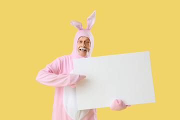 Senior man in Easter bunny costume pointing at blank poster on yellow background
