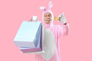 Senior man in Easter bunny costume with credit card and shopping bags on pink background
