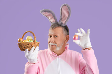 Senior man in bunny costume holding wicker basket with Easter eggs on lilac background