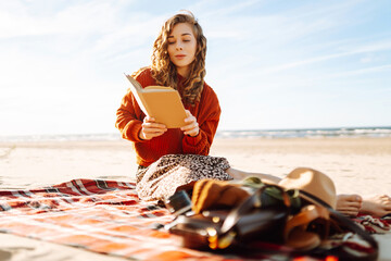 Beautiful woman sitting on the blanket reading a book at the beach picnic. The concept of relaxation, enjoyment, solitude with nature.