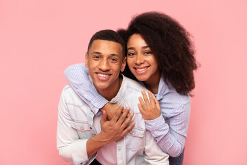 Lovely young black woman hugging her husband from behind, feeling happy on pink studio background,...