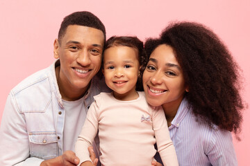 Fototapeta premium Portrait of happy black parents and their little daughter smiling to camera, family of three sharing moment of love and happiness, pink background