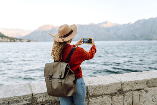 Female traveler in casual sweater taking picture of amazing landscape with mountains, ocean and sunset sky on smartphone during vacation.Tourist during holidays.