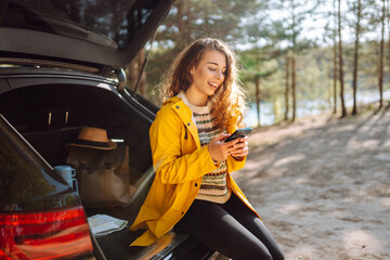 Young woman sitting in open trunk with phone. Lifestyle, travel, tourism, nature, active life.