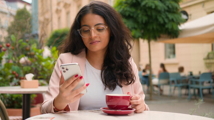 Successful Indian Arabian woman ethnic female girl businesswoman looking mobile phone drinking cup coffee morning breakfast city street cafe restaurant outside. Business travelling relaxation weekend