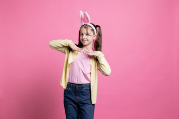 Adorable kid wearing bunny ears for easter photoshoot in studio, celebrating spring holiday over pink background. Cute little girl being playful and festive for traditional celebration.