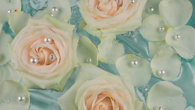 Pearls fall on a luxurious light blue background of flowers and white rose petals floating on the surface of the water. Slow motion.