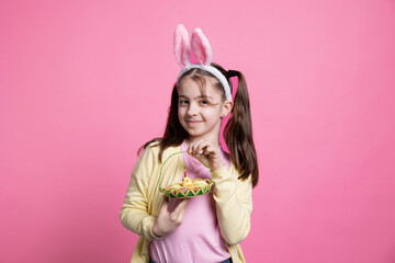 Young joyful child posing with easter eggs and toys on camera, feeling happy and excited about spring holiday celebration and gifts. Little girl with bunny ears smiling in studio.
