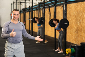 Fototapeta na wymiar Positive energetic middle-aged male fitness instructor standing with thumbs-up gesture in gym, with people exercising in blurred background