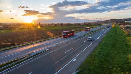 Two red buses  drive at high speed on the highway through the rural landscape. Fast blurred highway driving. A scene of speeding on the highway. Beautiful sunset in the background.