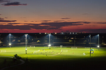Aerial view of public sports park with people engaged in football game on grass stadium at sunset....