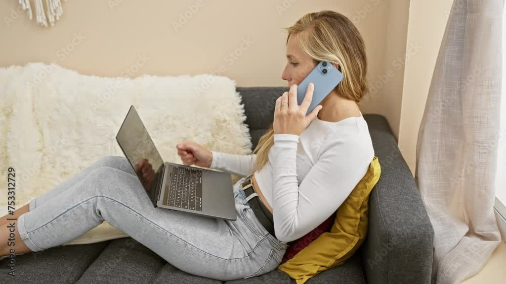 Wall mural A young woman multitasks with a phone and laptop in a cozy living room, portraying connectivity and modern home life.