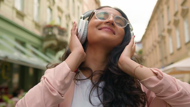 Joyful relaxed Indian Arabian ethnic teen gen z woman student lady female tourist enjoying listening music playlist app dancing song wireless headphones sound rhythm lifestyle in city street outdoors