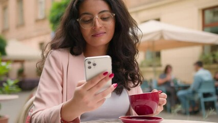 Close up thoughtful Indian Arabian woman ethnic female girl businesswoman tourist using looking mobile phone scrolling smartphone reading news social media holding coffee cup city street cafe outside