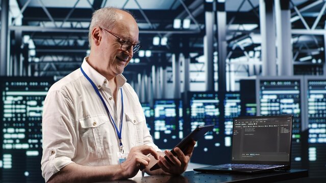 Old system administrator in data center, solving tasks on tablet while running scripts on laptop terminal. Qualified employee amidst servers, accessing databases and manipulating lines of code