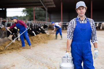 Portrait of adult man, dairy farmworker in uniform carrying metal milk can on farm in spring