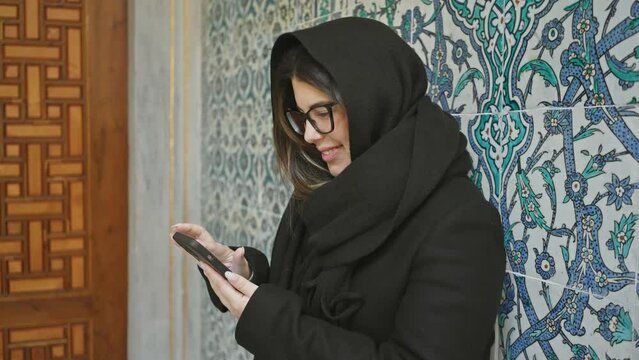 Smiling woman in hijab using smartphone with traditional turkish tiles in background, istanbul topkapi palace.