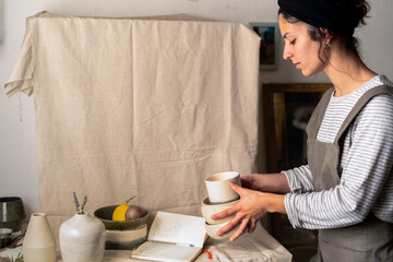 A ceramic artist holds her handmade pottery while checking stock in her notebook amidst a collection of unique ceramic creations