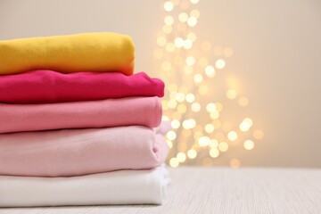 Stack of folded clothes on wooden table against beige background with blurred lights, space for text