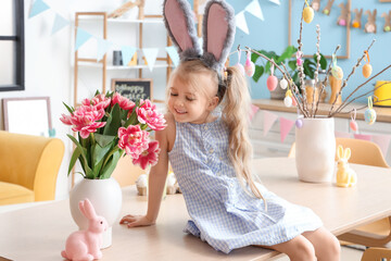 Cute little girl in bunny ears sitting on table with tulip flowers at home. Easter celebration