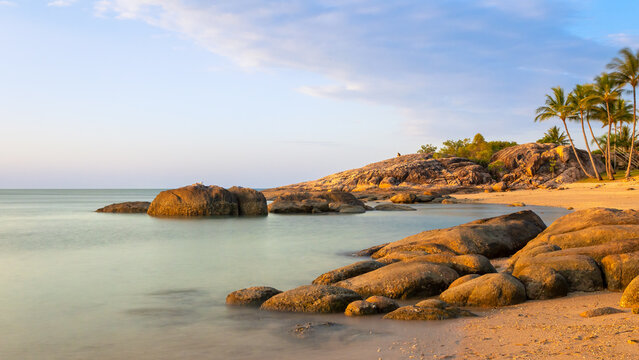 An idyllic tropical beach with smooth water, palm trees, granite rock formations and blue sky and clouds in the town of Bowen in the Whitsunday Great Barrier reef region of Queensland, Australia.